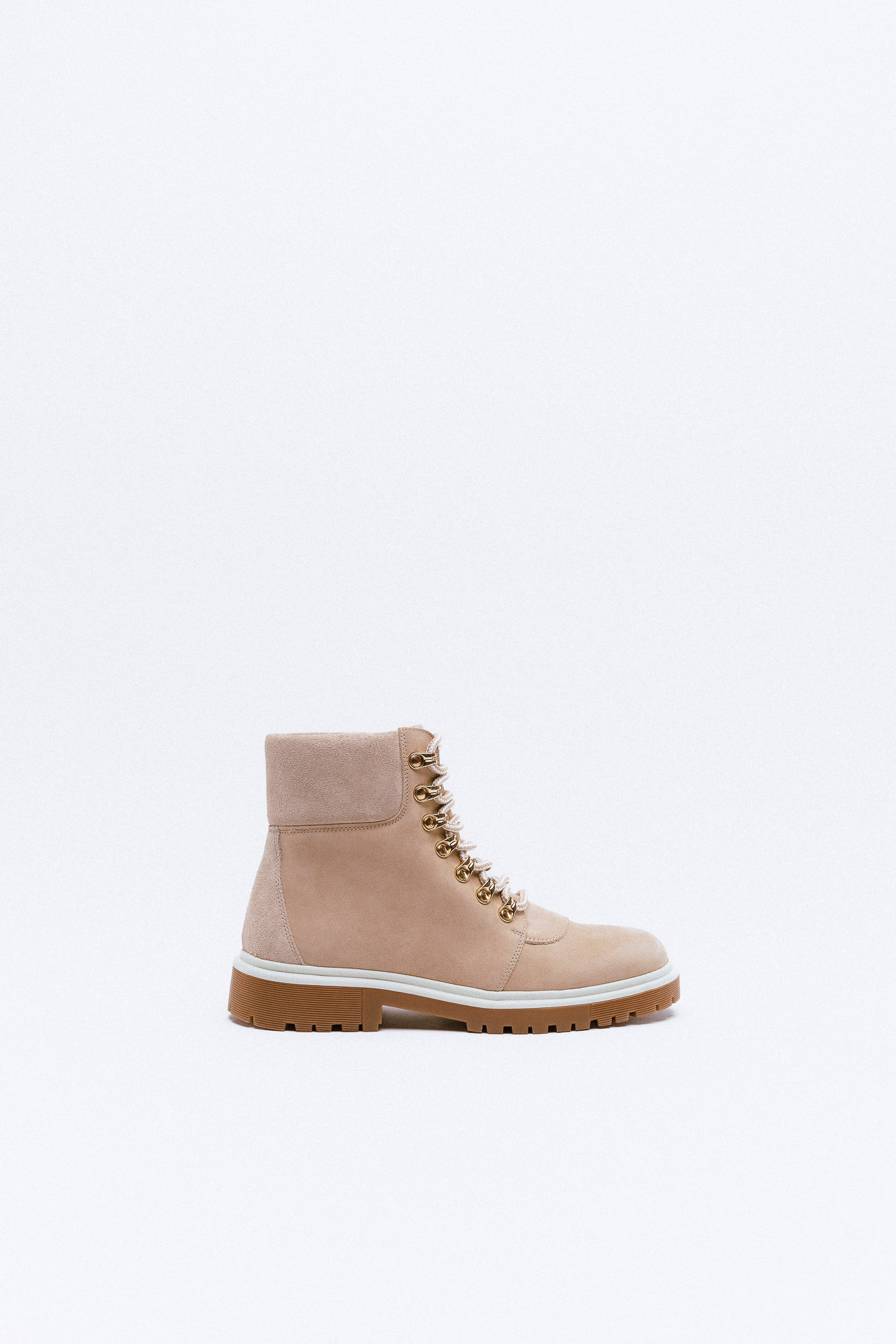 A beige suede combat boot with gold hardware, featuring a chunky brown sole and white trim.
